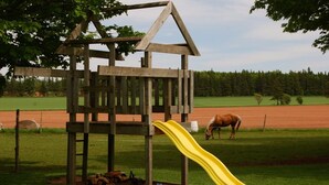 A wooden playhouse with a yellow slide in a grassy field. - Bugaboo Cottages (Central Bedeque)