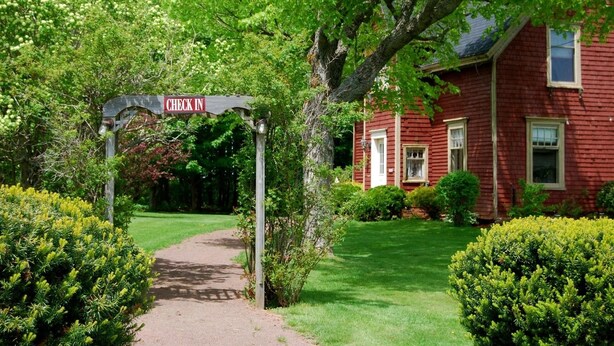 A red wooden house with a 'CHECK IN' sign in front, surrounded by lush greenery and a well-maintained lawn. - Bugaboo Cottages (Central Bedeque)