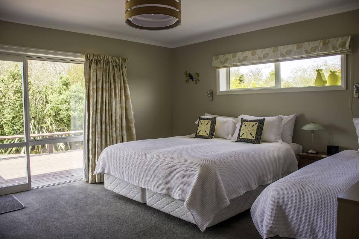 A bedroom with two beds, a window with a view of greenery, a curtain, and a bedside table with a lamp.