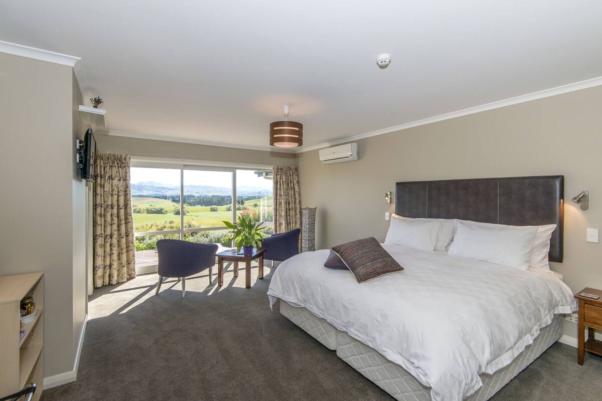 A bedroom with a large bed, a desk, and a chair. There is a window with curtains and a view of a green landscape.