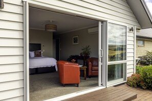 A room with a bed, a chair, a table, and a sliding glass door leading to an outdoor area with a wooden deck.