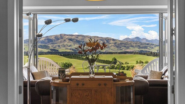 A room with a view of rolling hills and mountains. There is a wooden table with a vase of flowers and a lamp.