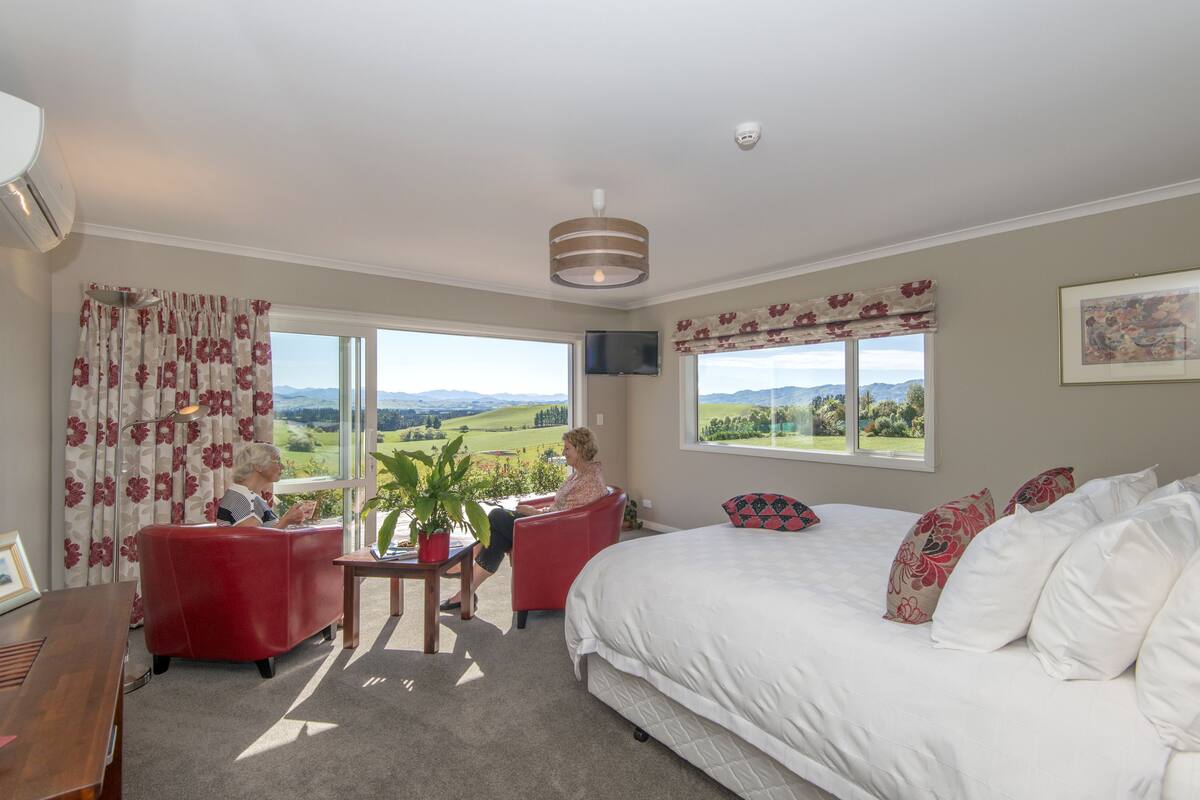 A bedroom with a large bed, a red armchair, a small table with a plant, and a view of the countryside through large windows.