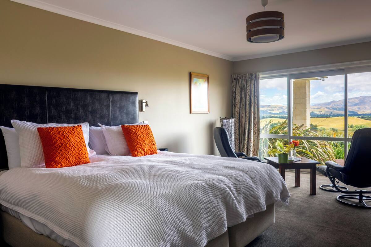 A hotel room with a large bed, a view of the countryside, a desk, and a chair.