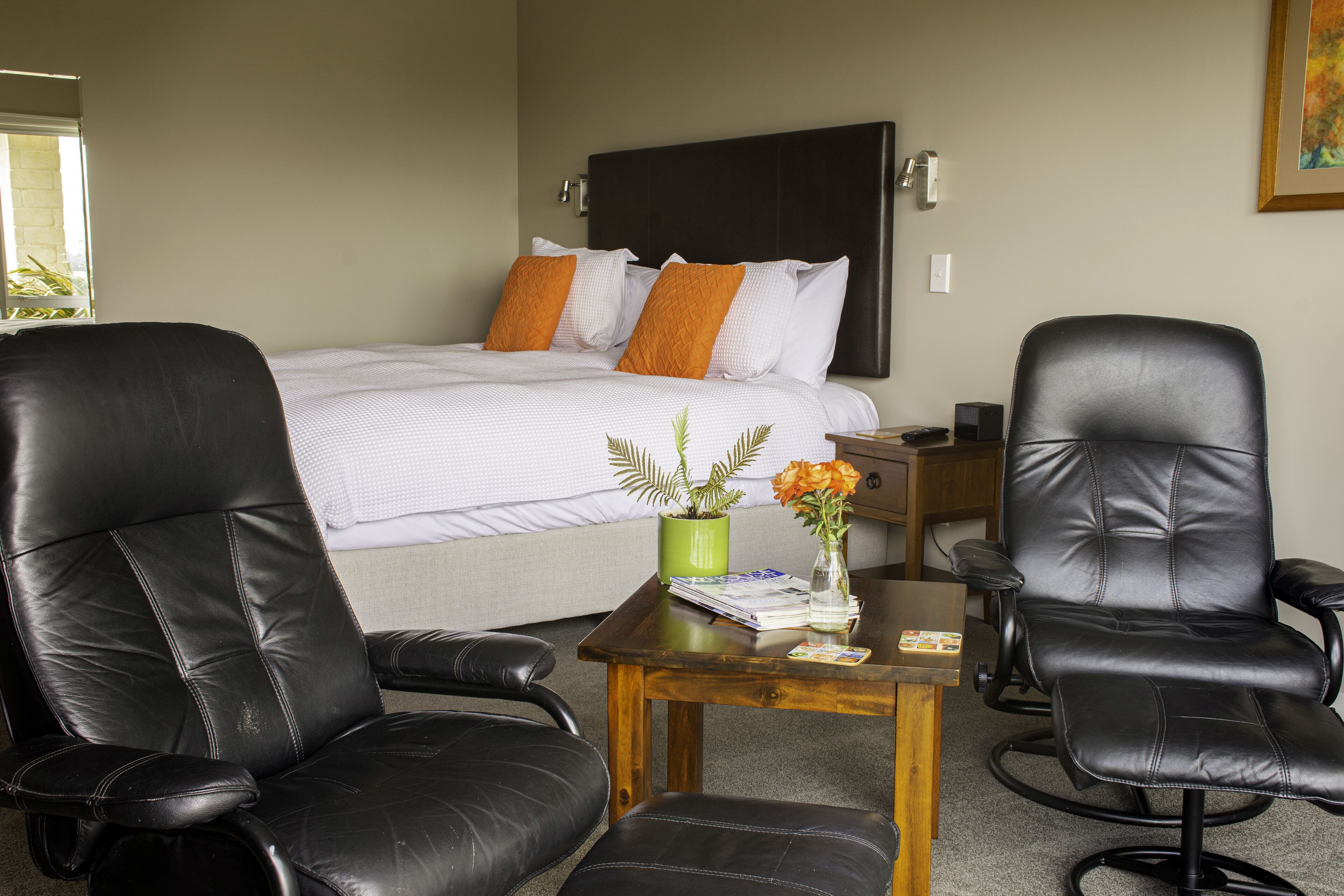 Quarto de hotel com uma cama, duas poltronas de couro preto, uma mesinha de madeira com um vaso de flores e uma planta verde.