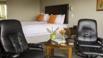 Quarto de hotel com uma cama, duas poltronas de couro preto, uma mesinha de madeira com um vaso de flores e uma planta verde.