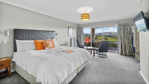 A modern hotel room with a large bed, a desk, and a chair. There is a television on the wall and a window with a view of the countryside.