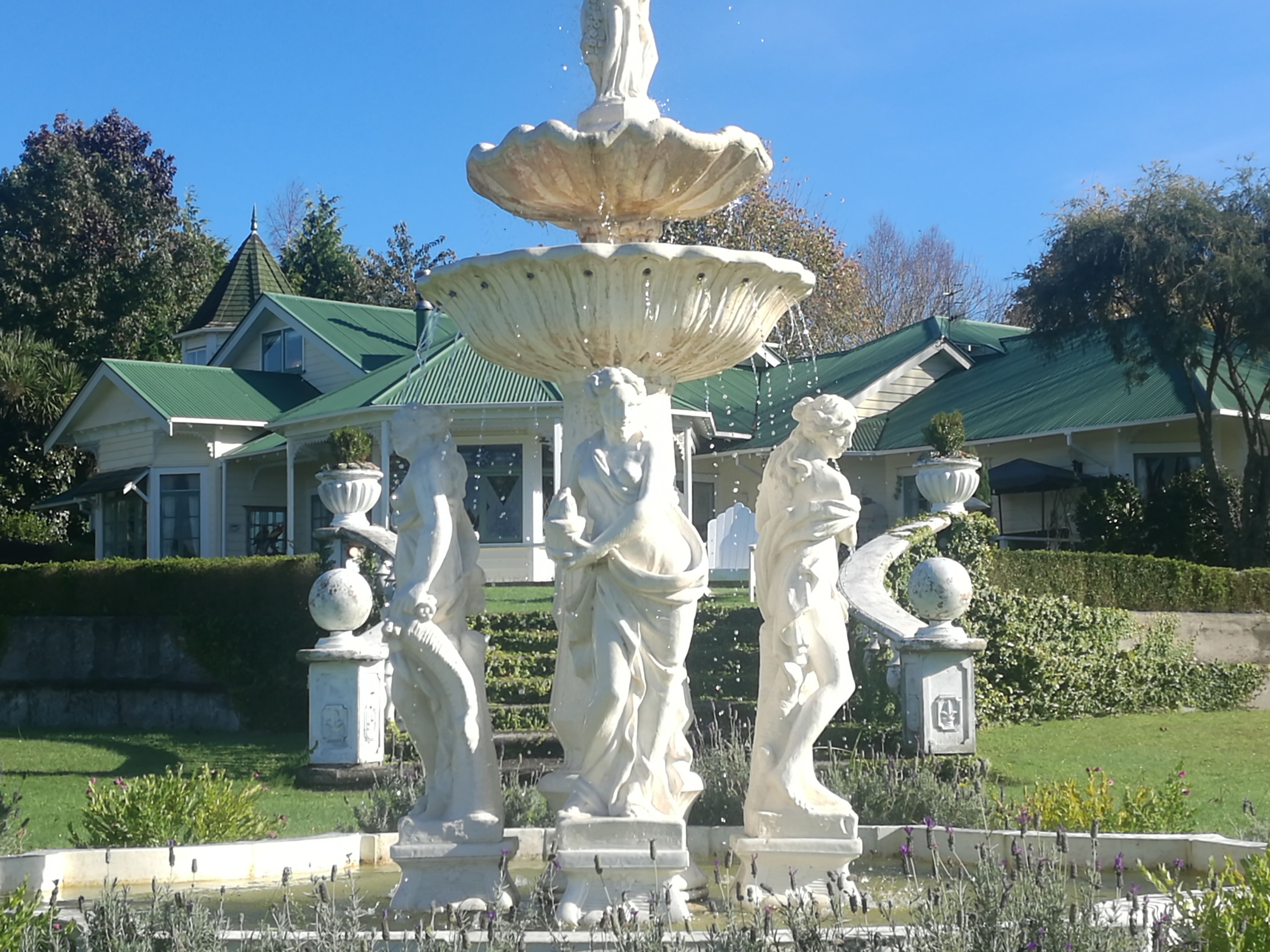 A fountain with statues in a garden area, surrounded by a well-maintained lawn and a building with a green roof in the background.
