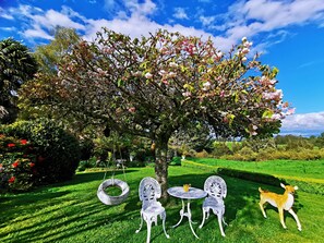 A garden with a tree, two white chairs, a small table, and a dog. - Country Villa Estate (Hamurana)