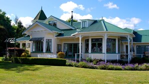 A large, two-story house with a green roof and white exterior. It features a covered porch with a railing and a small garden with purple flowers in front. - Country Villa Estate (Hamurana)