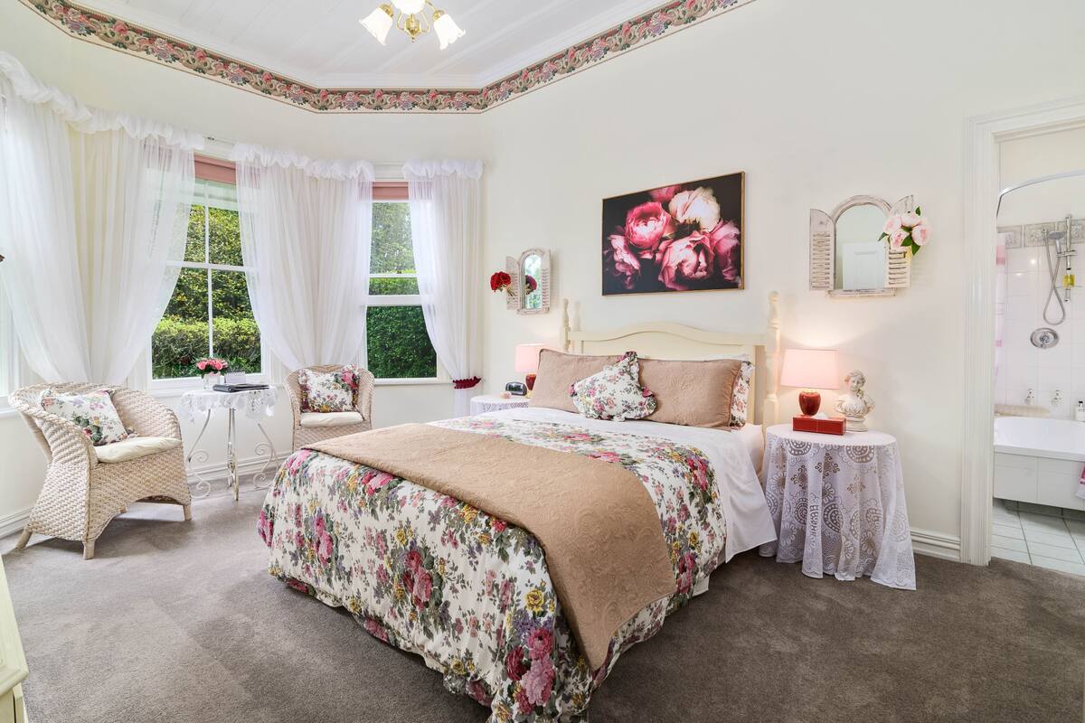 A bedroom with a large bed, a sitting area with a chair, a small table, and a bathroom with a sink and shower.