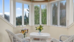 A sunroom with white wicker furniture, a central table with flowers, and large windows with a view of trees. - Country Villa Estate (Hamurana)