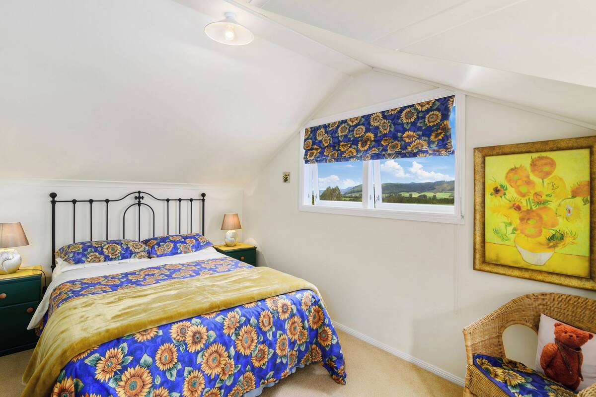A bedroom with a bed, nightstands, a window with a sunflower-patterned valance, a wicker chair, and a painting on the wall.