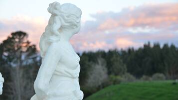 Una estatua blanca de una mujer con una flor en el cabello, con un cielo de atardecer y un bosque como fondo.