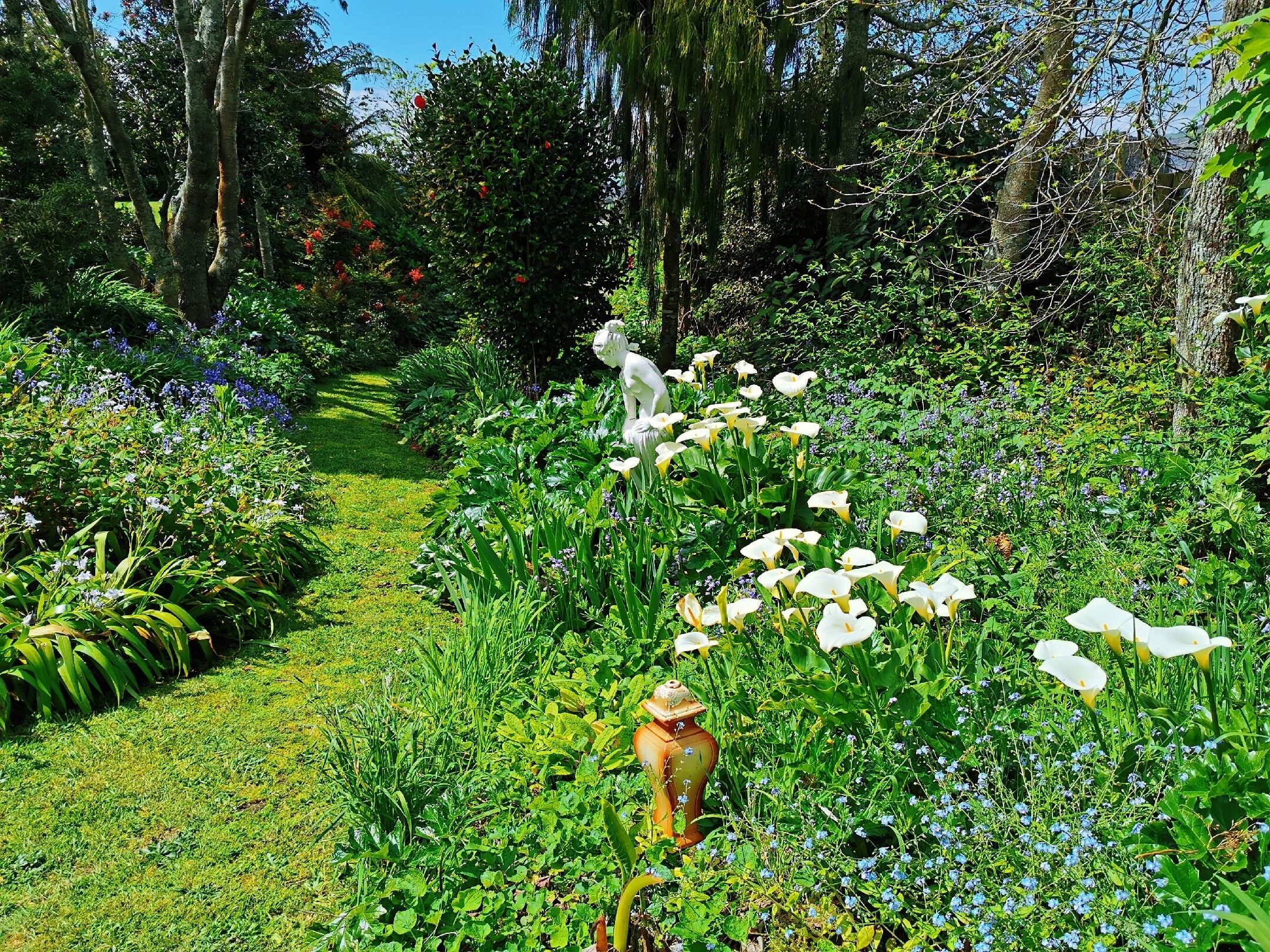 Un jardín con una estatua blanca de un ave, un jarrón amarillo y diversas plantas y flores.