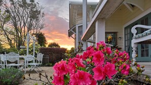 A garden with white metal chairs and tables, a hedge, and pink flowers. - Country Villa Estate (Hamurana)