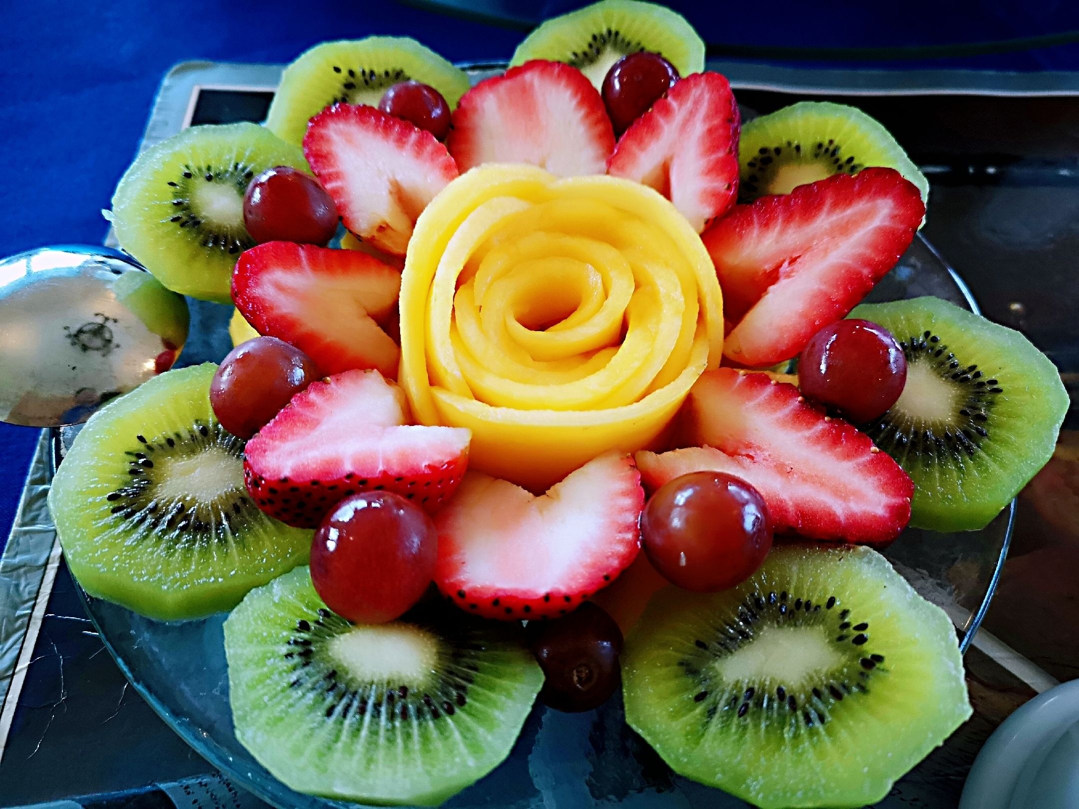Un plateau de fruits avec une composition centrale de fruits jaunes, entourée de fraises, de raisins et de kiwis coupés en tranches.
