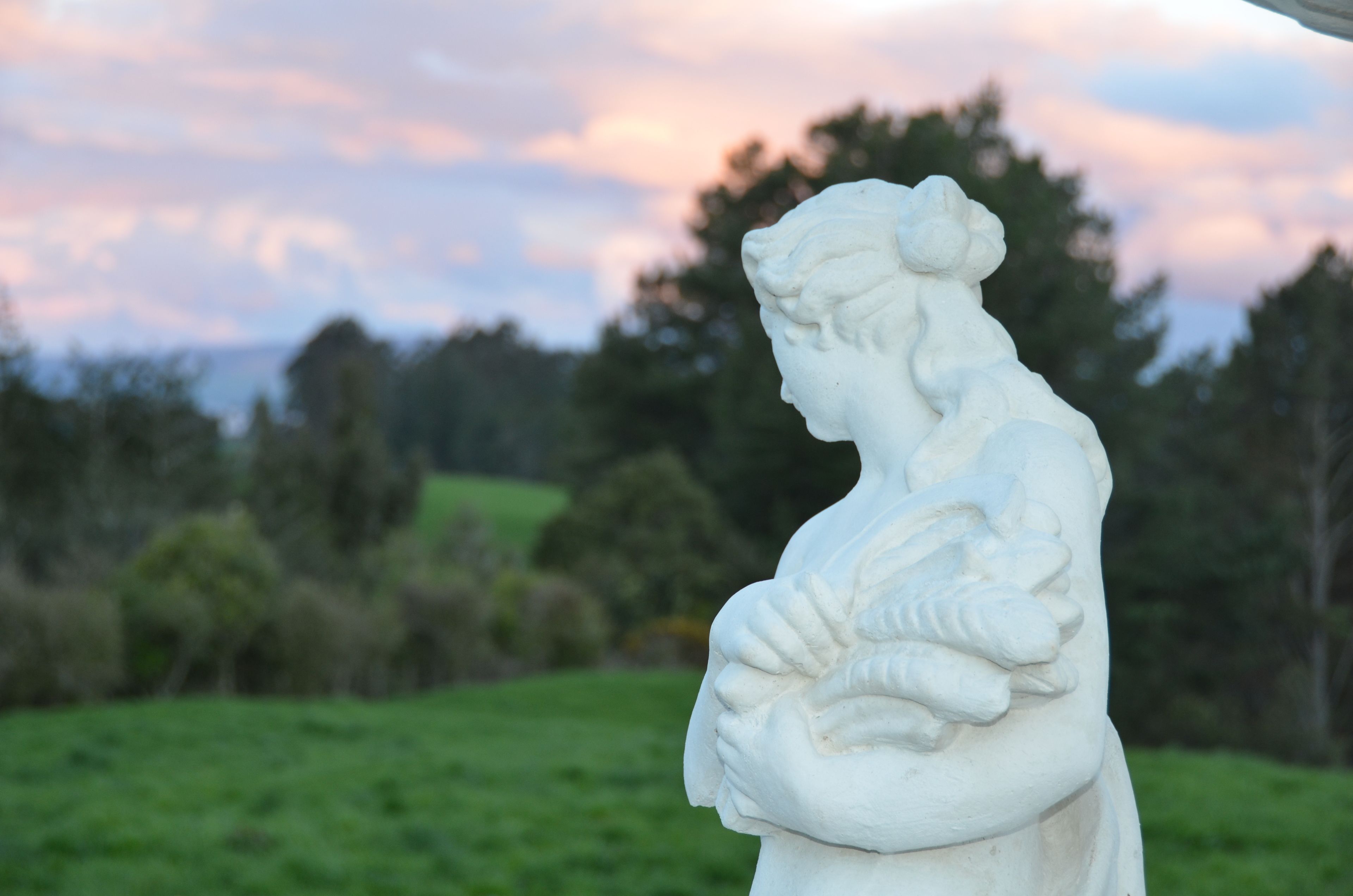 A white statue of a woman holding a child, set against a backdrop of trees and a cloudy sky.