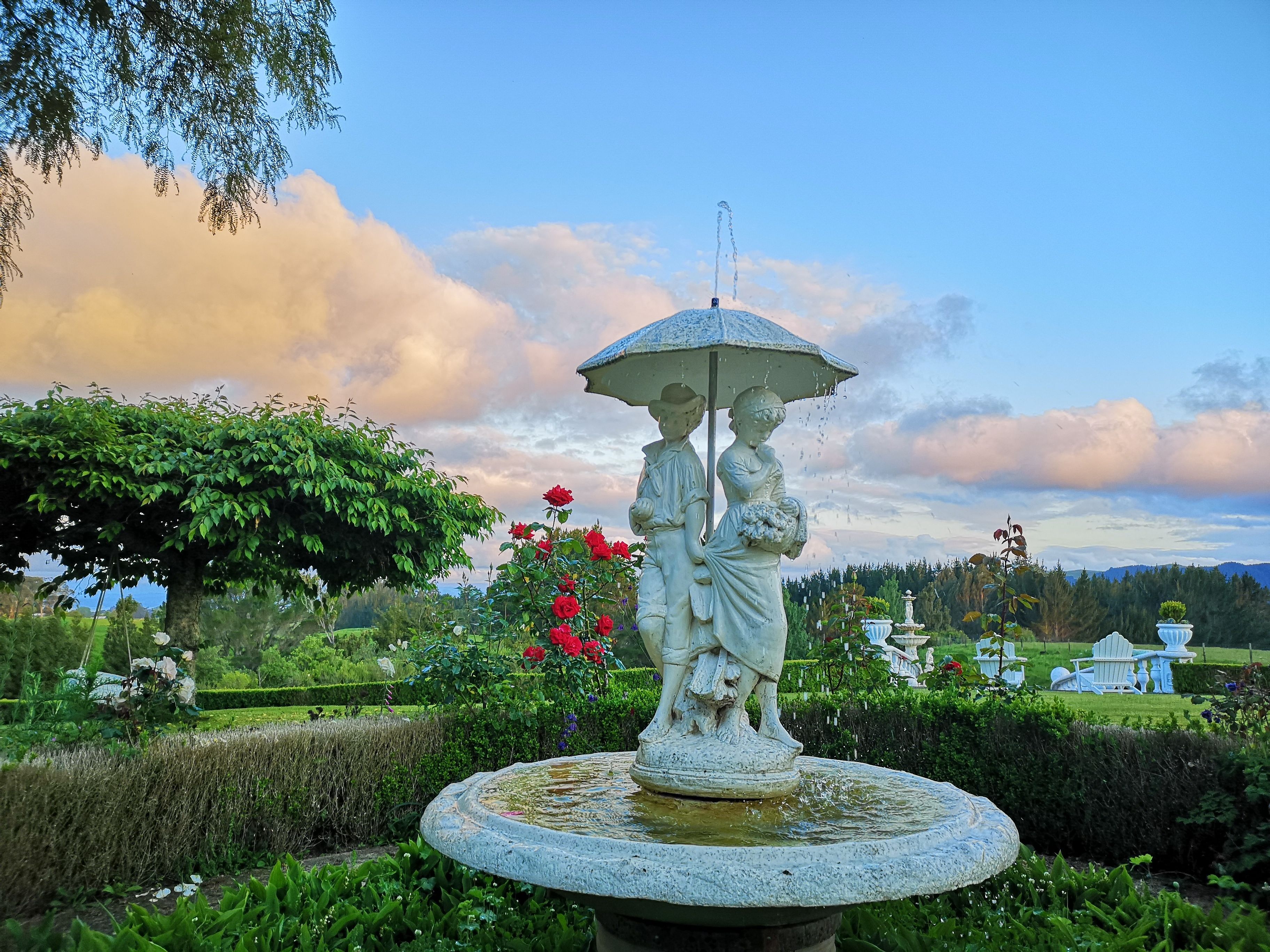 A garden with a statue of two figures under an umbrella, surrounded by greenery and flowers.