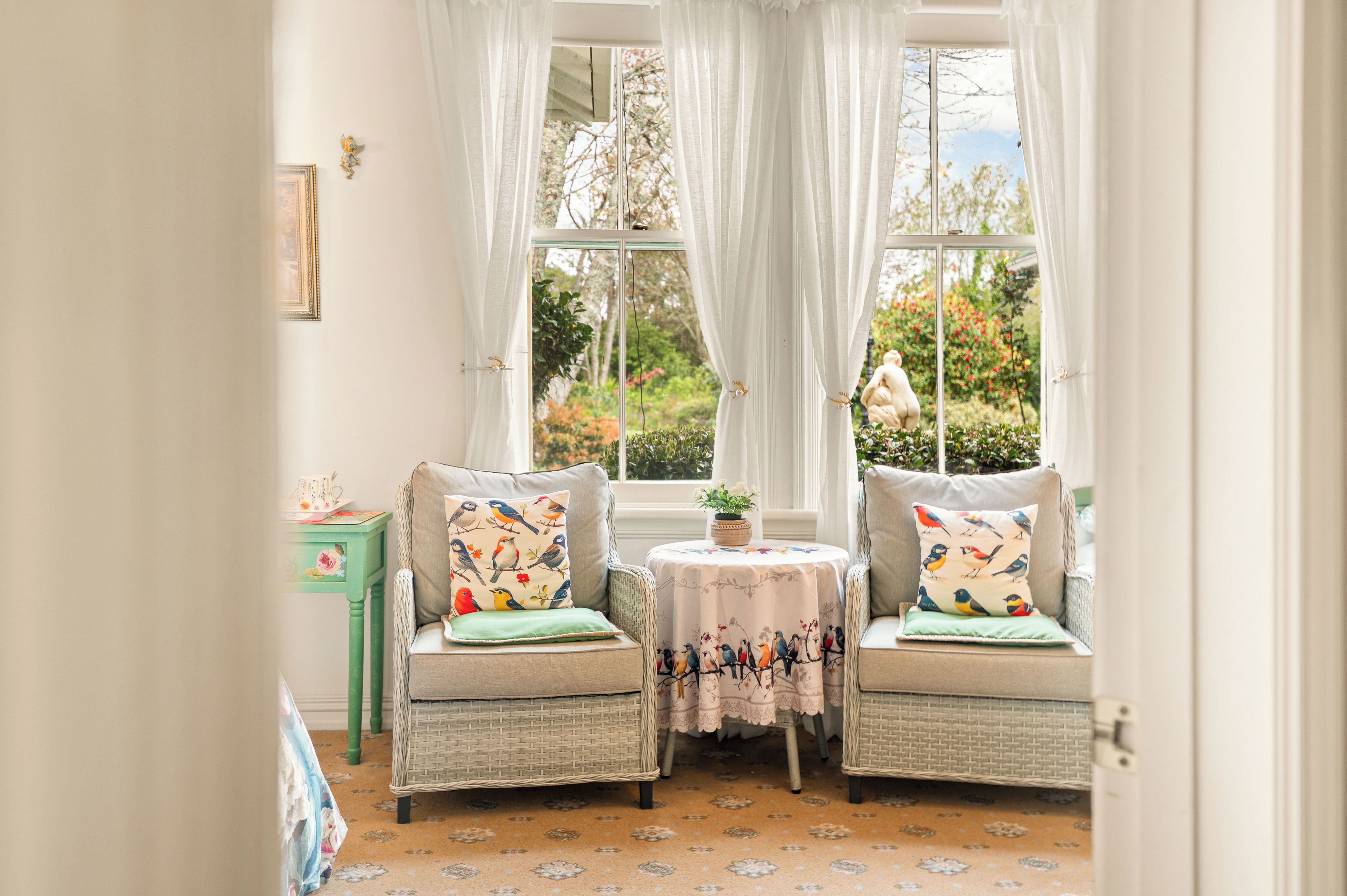 Ein Zimmer mit zwei Korbstühlen, einem kleinen Tisch mit einer blumigen Tischdecke und Blick auf einen Garten durch große Fenster mit weißen Vorhängen.