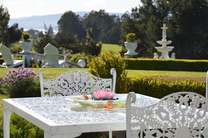 A white outdoor dining table with intricate metalwork, surrounded by white chairs, set in a lush garden with a fountain and neatly trimmed hedges in the background. - Country Villa Estate (Hamurana)