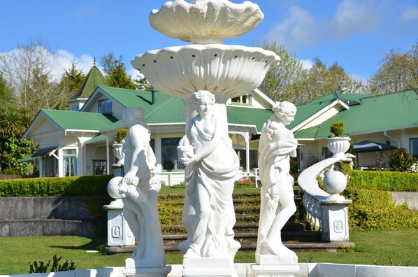 A white fountain with statues in a garden area, surrounded by a well-maintained lawn and a house with a green roof in the background. - Country Villa Estate (Hamurana)