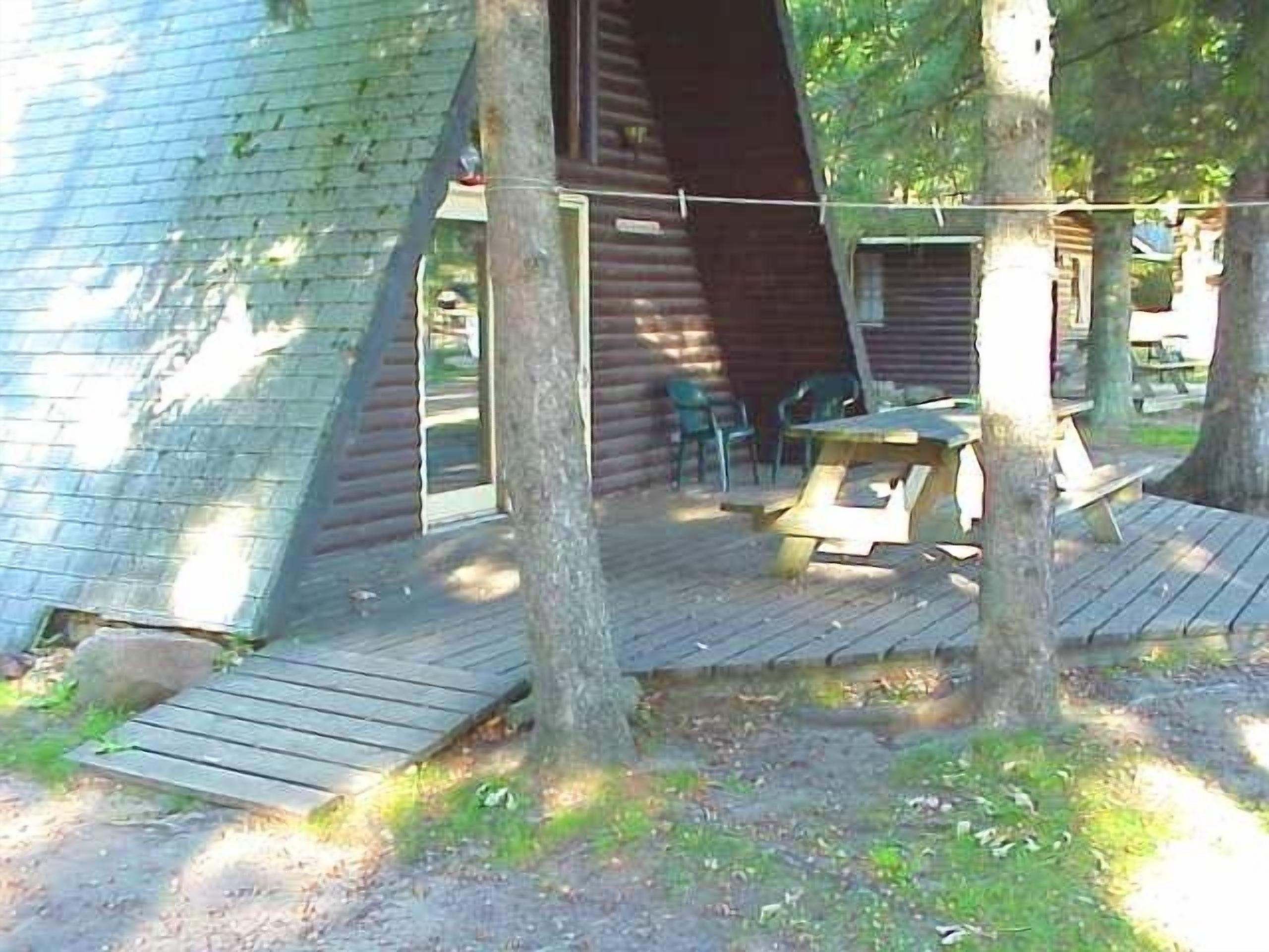 Une cabane en bois avec un toit en pente, entourée d’arbres. On y trouve une table de pique-nique et des chaises à l’extérieur.