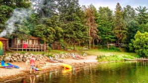 Una cabaña con chimenea humeante, rodeada de árboles y un lago.