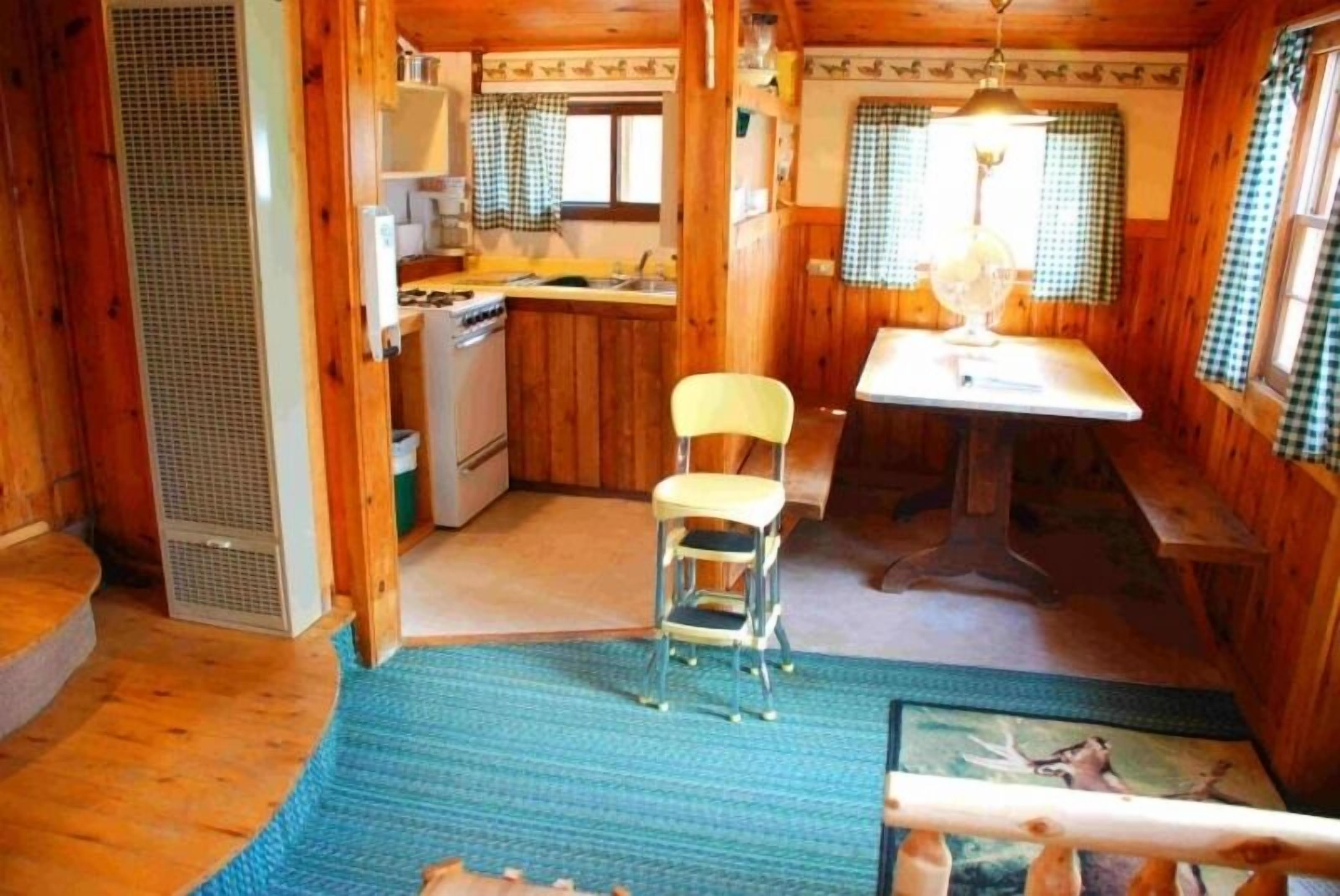 Eine kleine Küche mit Holzausstattung, einer Spüle und einem Herd. Ein gelber Stuhl steht in der Nähe der Spüle, und ein Tisch mit einer Lampe befindet sich im Vordergrund.