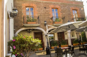 Un edificio de ladrillo con tres ventanas, cada una con balcón y macetas. Hay mesas y sillas exteriores, y una sombrilla blanca que da sombra.
