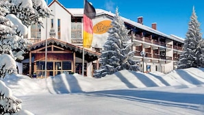 A hotel with a traditional wooden facade, a balcony with a view of the snow-covered landscape, and a flag indicating it is a ski resort. - Hotel St. Georg Gmbh (Bad Aibling)