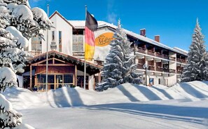Ein Hotel mit traditioneller Holzfassade, einem Balkon mit Blick auf die verschneite Landschaft und einer Flagge, die es als Skigebiet kennzeichnet.