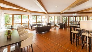A spacious room with a high ceiling, wooden beams, large windows, and a mix of seating including a sofa, chairs, and a bar area. - Hotel St. Georg Gmbh (Bad Aibling)