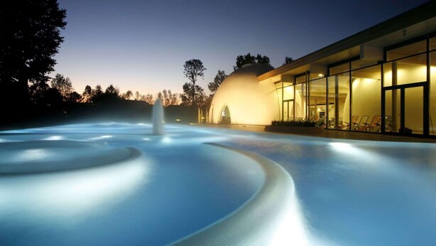 A swimming pool with a fountain, surrounded by a building with large windows and a dome structure. - Hotel St. Georg Gmbh (Bad Aibling)