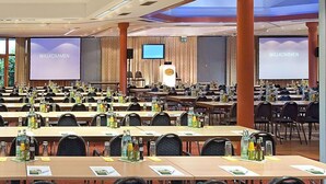 Un salón de conferencias con varias mesas, sillas y una pantalla grande que muestra “WILLKOMMEN”.