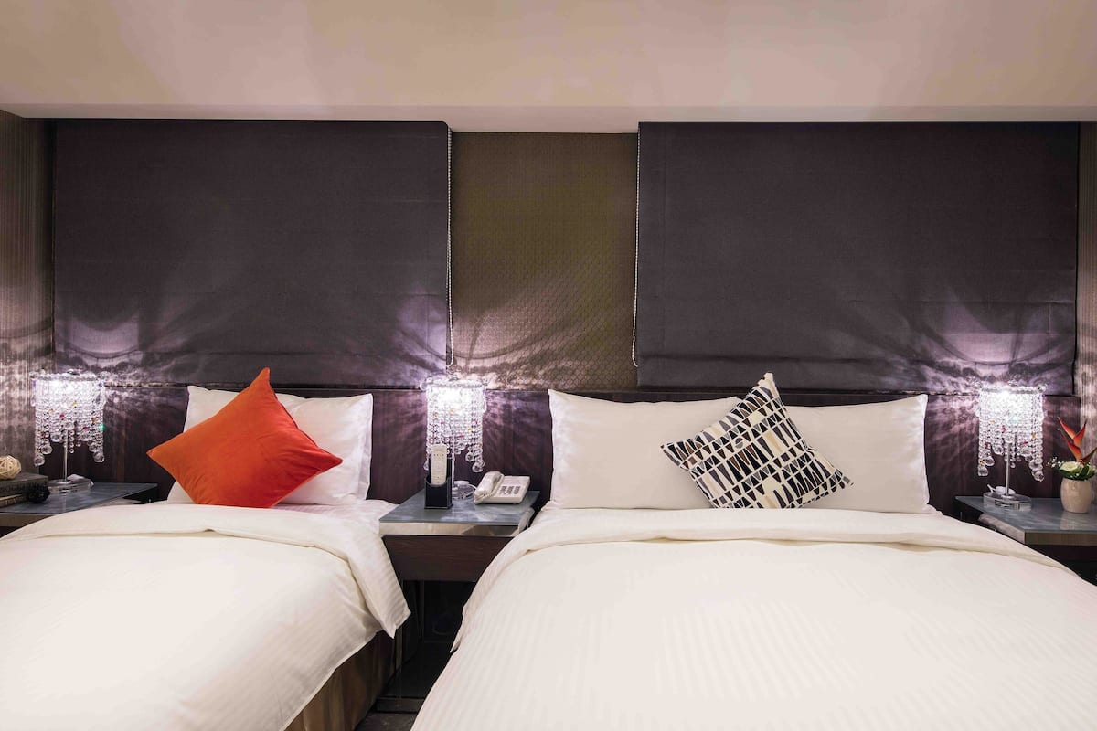 A hotel room with two beds, each with a white comforter and pillows, a nightstand with a phone and a lamp, and a wall-mounted light fixture.