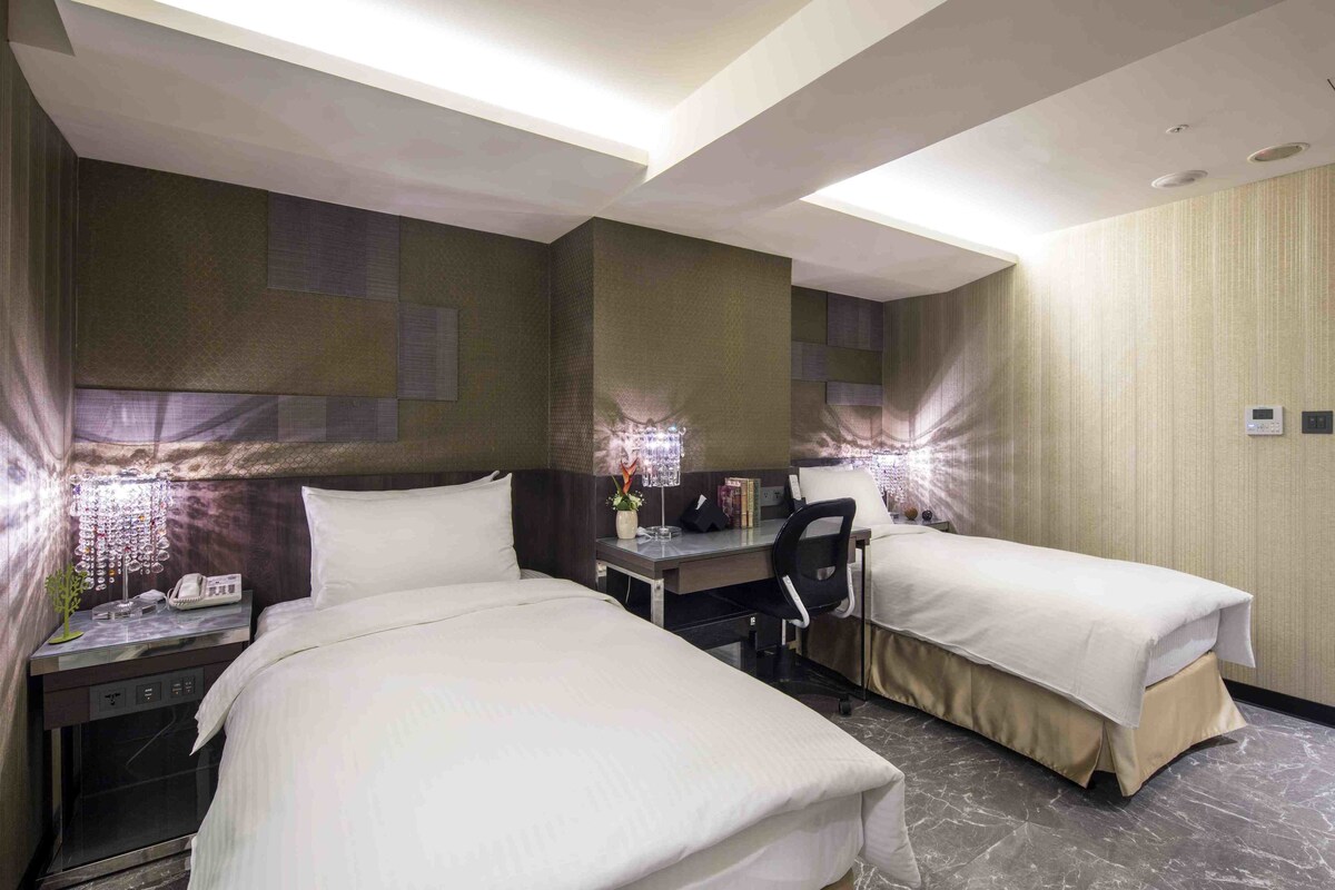 A hotel room with two beds, a desk, and a chair. There are lamps on the nightstands, a phone, and a small plant.