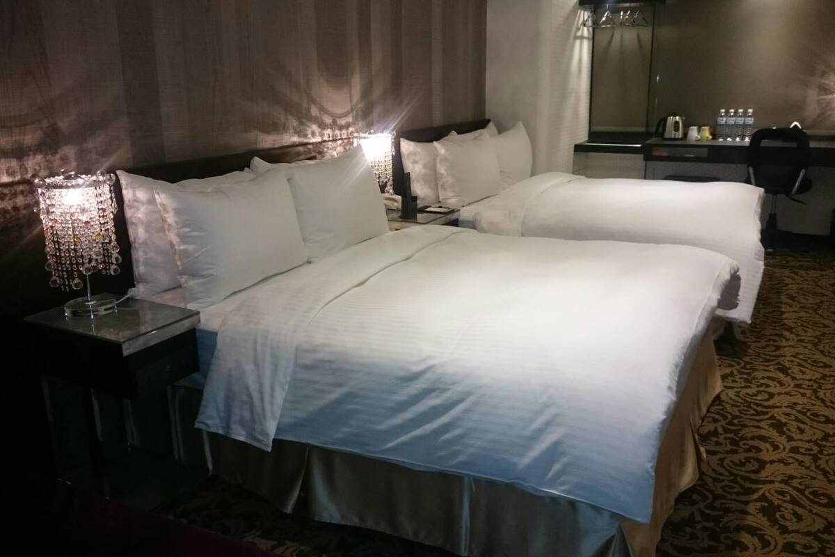 A hotel room with two beds, a nightstand with a lamp, a mirror, and a window with a city view.