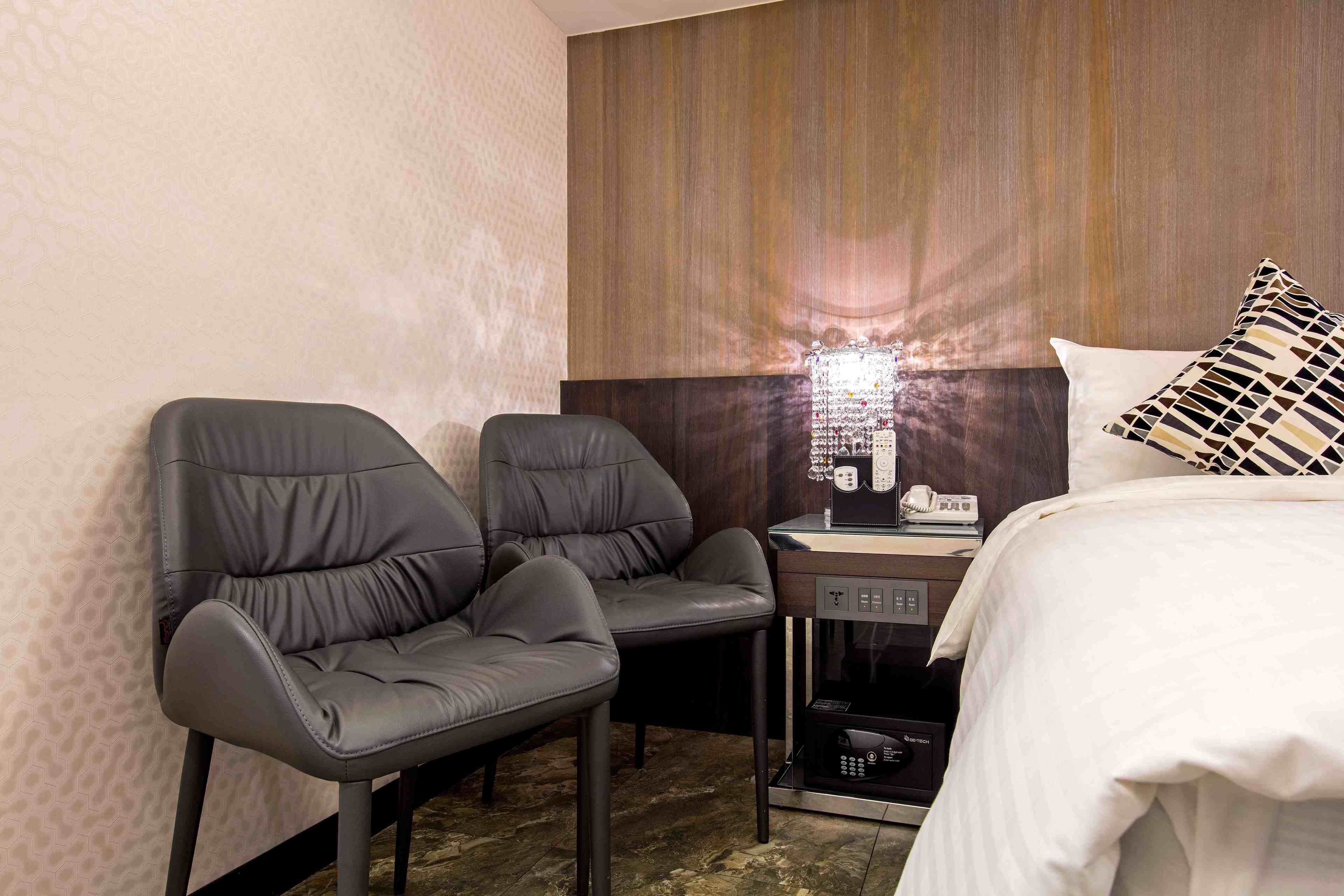 A hotel room with a bed, two chairs, a small table with a lamp, and a minibar.