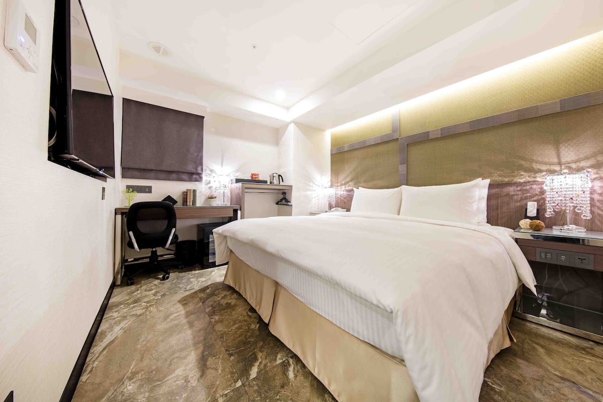 A hotel room with a large bed, a desk with a chair, a small kitchenette, and a mirror on the wall.