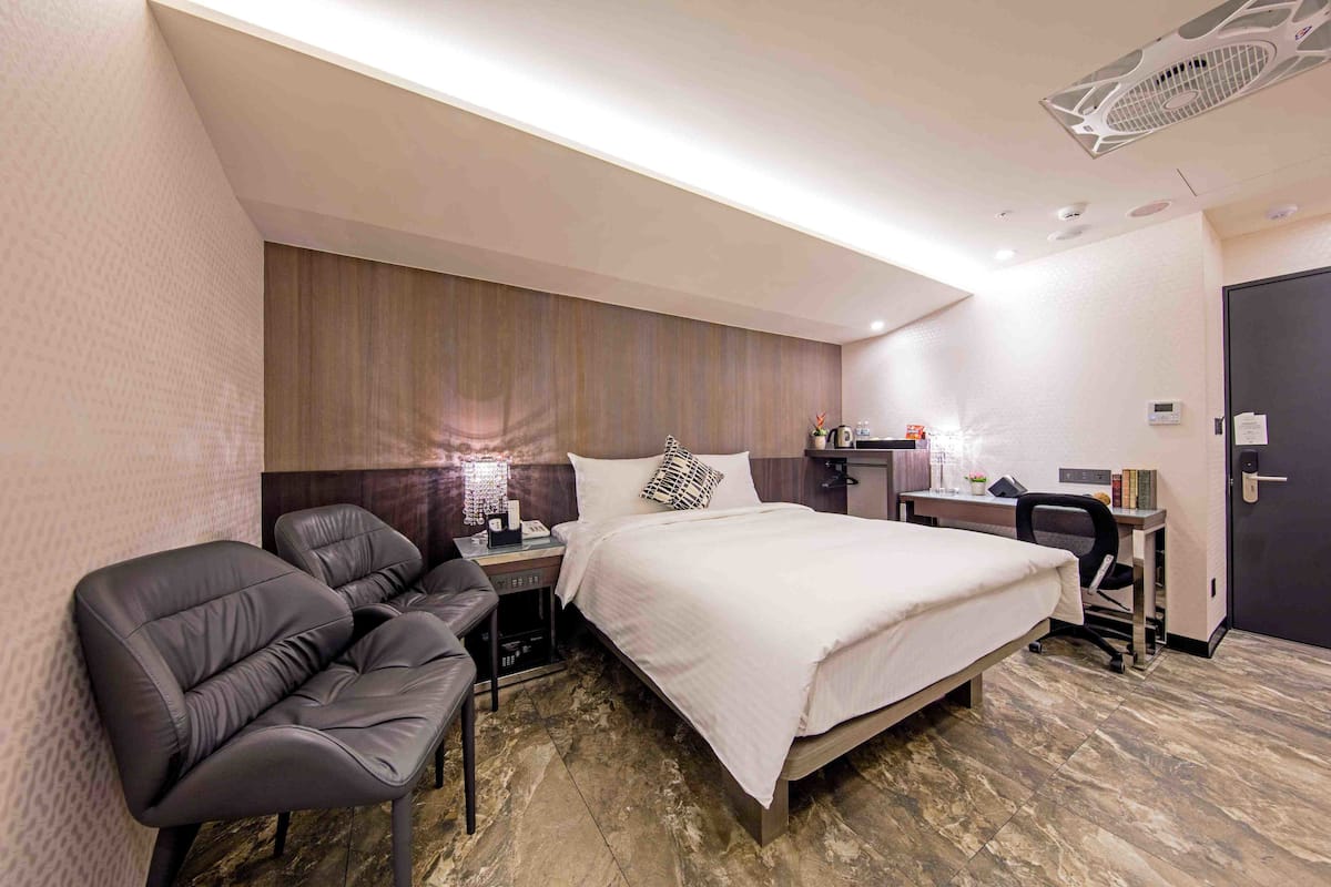 A modern hotel room with a large bed, a desk, a chair, and a small table with a lamp.