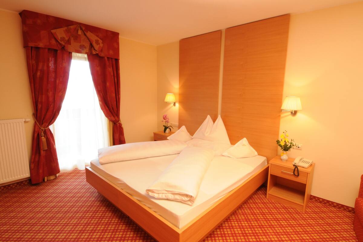 A hotel room with a large bed, two bedside tables with lamps, a wooden headboard, and a red checkered carpet.