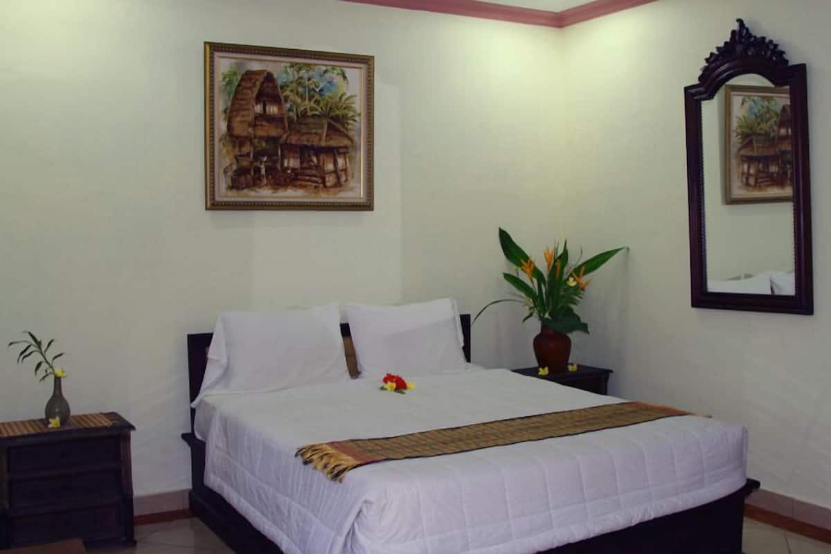 A neatly made bed with white linens, a bedside table with a vase and a plant, and a painting of a house on the wall.
