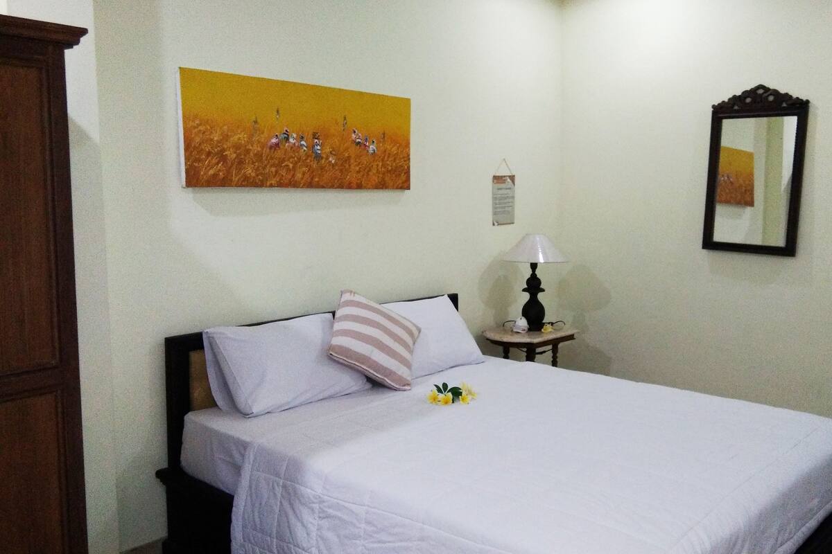 A neatly made bed with white linens, a striped pillow, and a small bouquet of yellow flowers. A lamp is on a bedside table, and a framed painting hangs on the wall.
