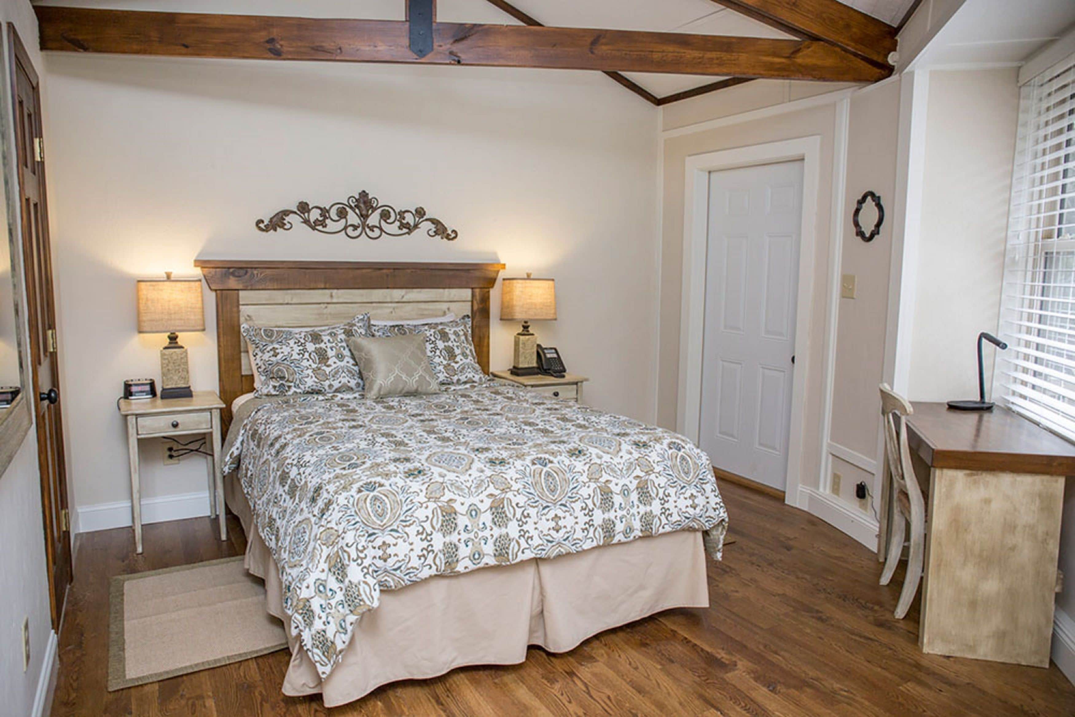 A bedroom with a wooden headboard, two bedside tables with lamps, a bed with patterned bedding, a wooden desk, and a door.
