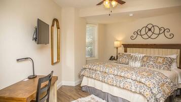 A bedroom with a bed, a desk, a chair, a ceiling fan, a TV, and a window with blinds.