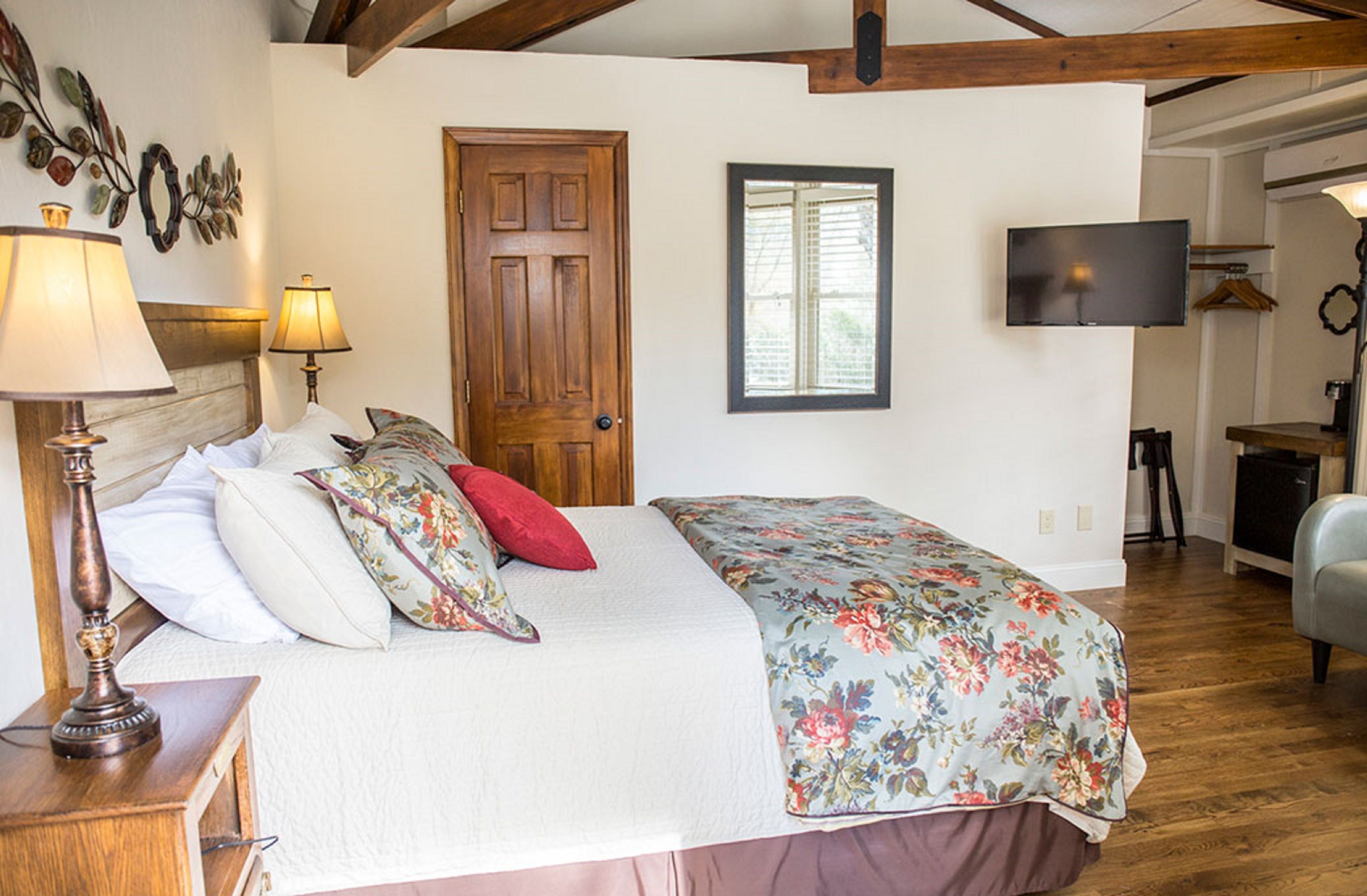 Un dormitorio con cabecera de madera, una cama con ropa de cama floral, una mesita de noche con lámpara, una puerta de madera, una ventana y un televisor montado en la pared.