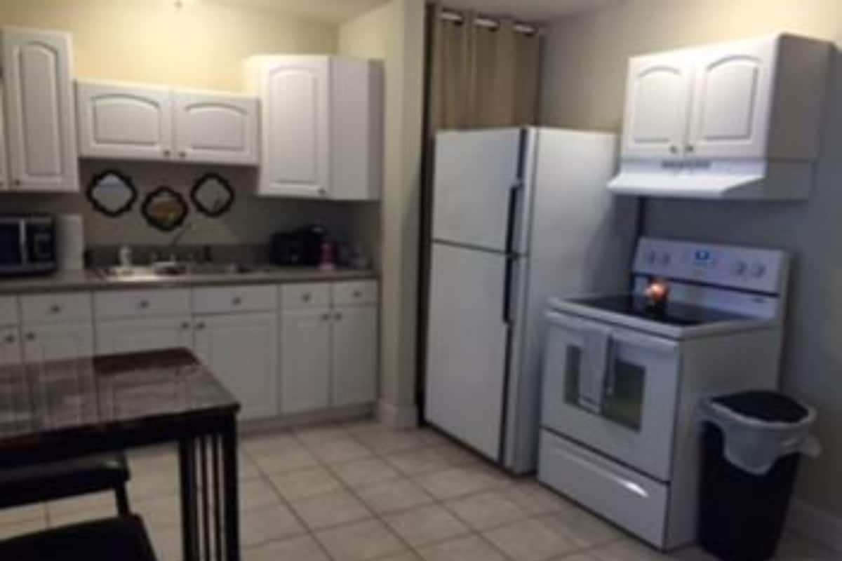 A compact kitchen with white cabinets, a stove, a refrigerator, and a trash bin.