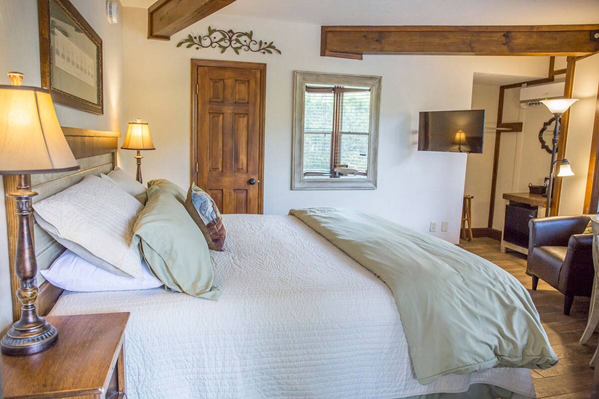 A bedroom with a large bed, two bedside lamps, a wooden door, a window, a TV, and a chair.