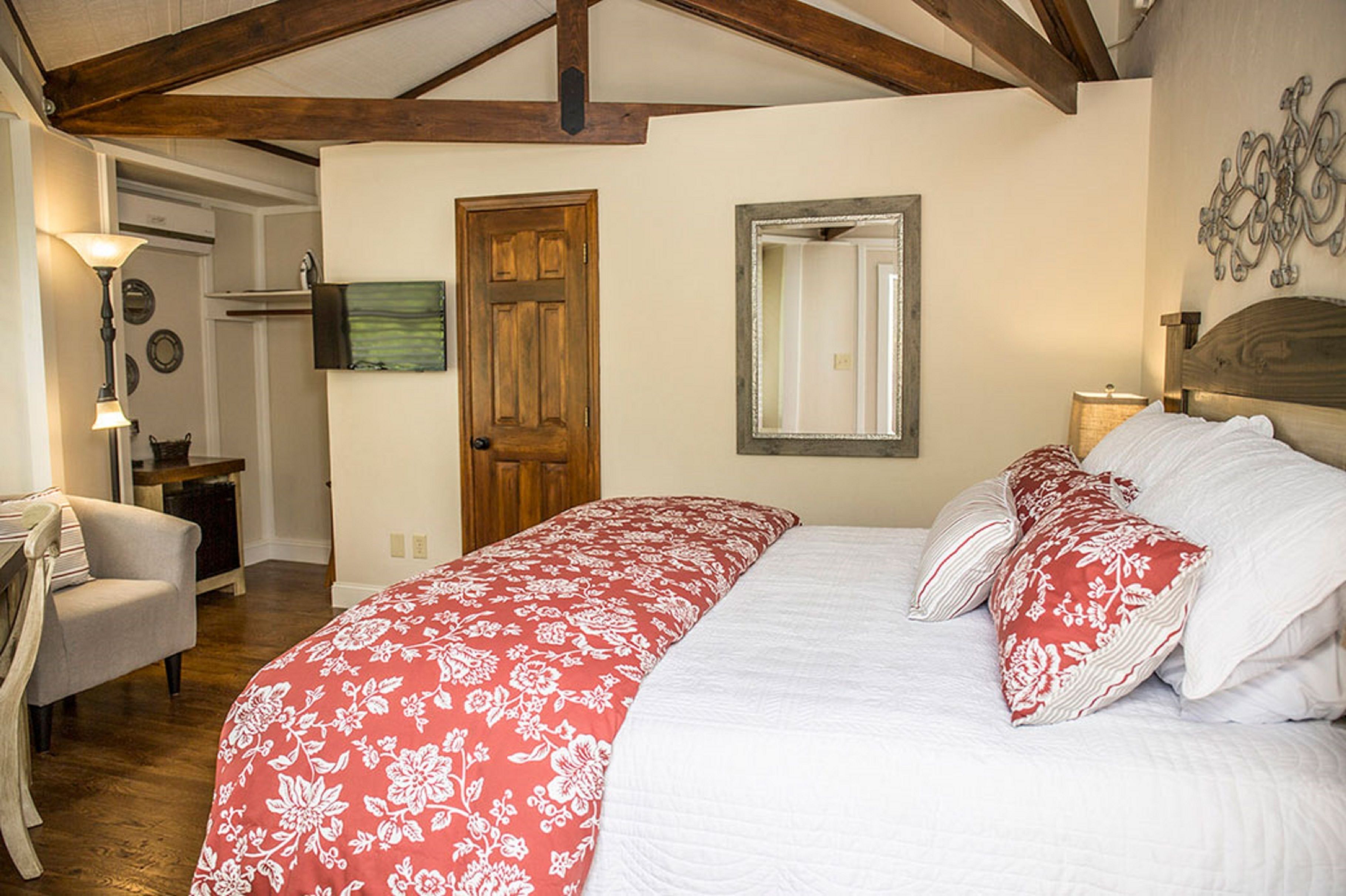 A bedroom with a large bed, wooden beams on the ceiling, a wooden door, a mirror, and a chair.